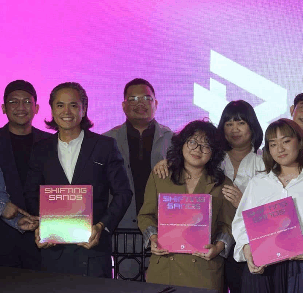 Team holding Shifting Sands branding project with bright pink background, showcasing brand identity work.