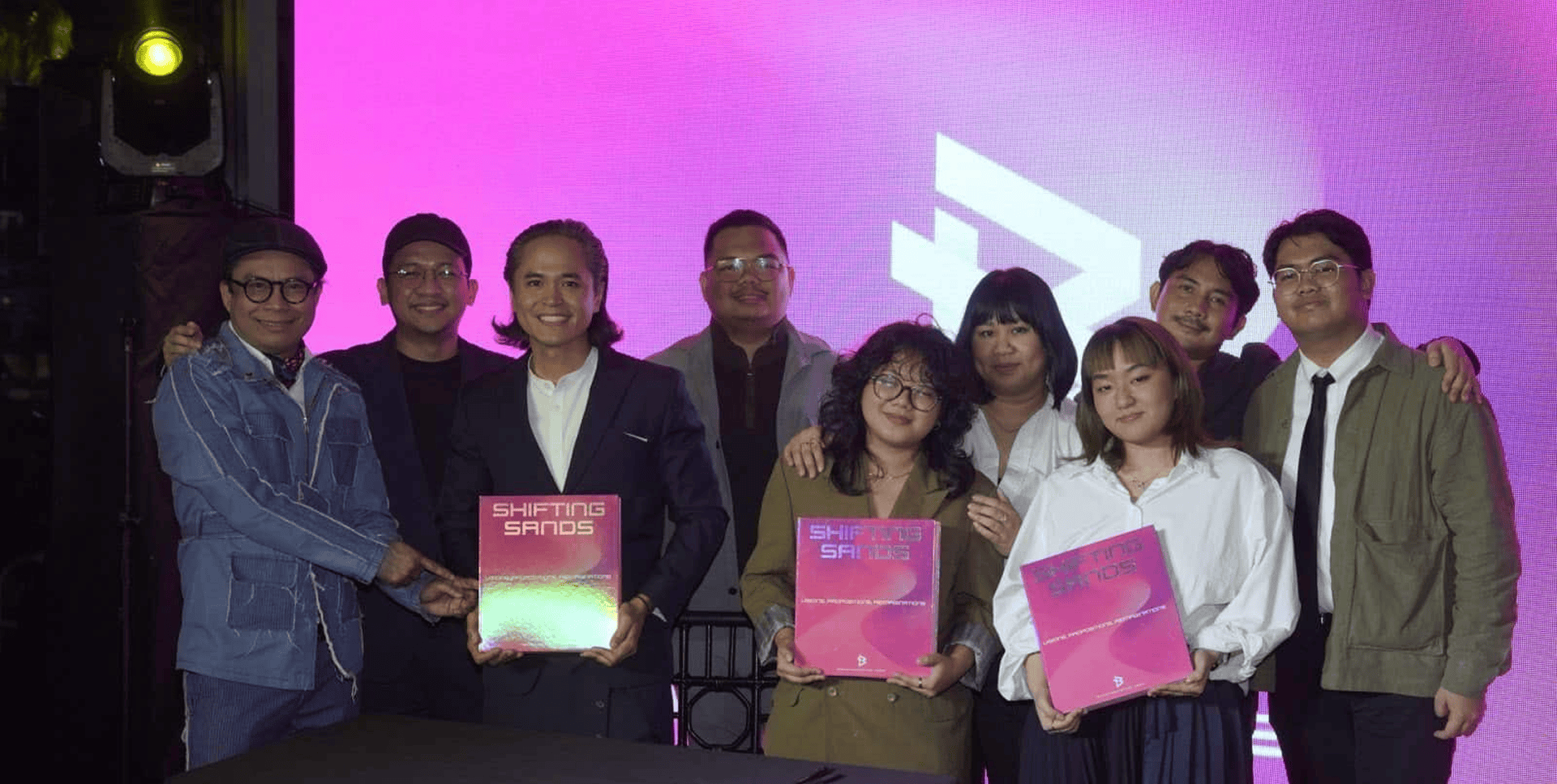 Team holding Shifting Sands branding project with bright pink background, showcasing brand identity work.
