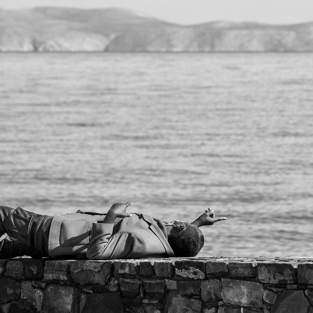 Person relaxing on a stone wall by the sea with mountains in the background. Peaceful outdoor scene.